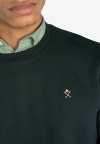 Sudadera verde oscura con cuello redondo, que presenta un pequeño logotipo rosa de herramientas cruzadas en el pecho, sobre una camisa de color verde claro con cuello.