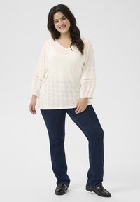 Crème blouse met perforaties, v-hals en balloonmouwen, gecombineerd met donkerblauwe rechttoe rechtaan jeans en zwarte instappers.