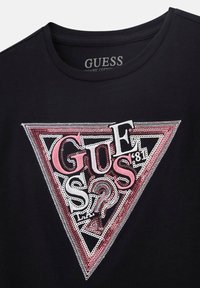 Černé tričko z organické bavlny s trojúhelníkovou grafikou v růžové a stříbrné barvě, se stylizovaným logem "Guess" a dekoračními flitry.