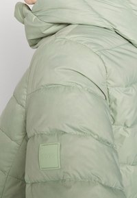 Gros plan sur une personne portant une veste d'hiver matelassée vert clair avec une capuche et un écusson carré de la marque sur la manche.