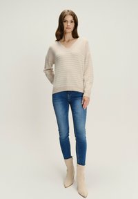 Lichtbeige gebreide trui met een V-hals en horizontale strepen, gecombineerd met blauwe skinny jeans en crèmekleurige enkellaarsjes. Glad textuur.