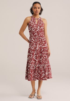 Naine kannab punast varrukateta midi kleiti valge abstraktse lillemustriga ja võtmegaelusega, seisab ühevärvilise tausta ees.