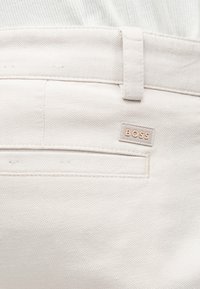 Gros plan d'un pantalon en tissu beige clair texturé montrant la poche arrière, la boucle de ceinture et une petite étiquette avec le logo BOSS au-dessus de la poche.