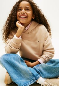 Niño sonriente con cabello rizado sentado con las piernas cruzadas, vistiendo un sudadera beige, vaqueros azules, calcetines blancos y zapatillas, contra un fondo liso.