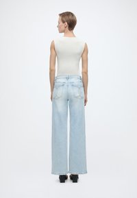 Άτομο όρθιο με πλάτη προς τα πίσω, φοράει άσπρο αμάνικο τοπ, ανοιχτό μπλε τζιν με φαρδιά μπατζάκια και ραμμένες πίσω τσέπες, και μαύρα σανδάλια με τακούνι.