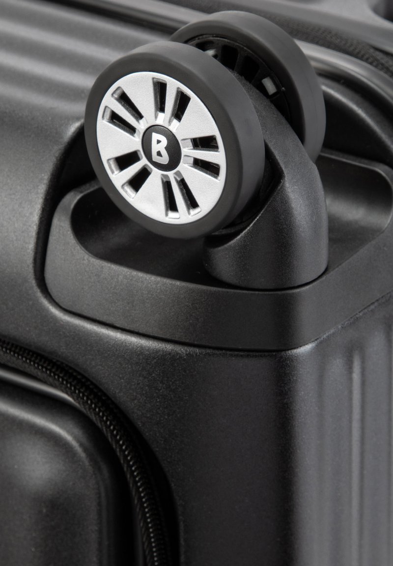 Eck eines schwarzen Hartschalenkoffers mit einem silbernen Rad, das ein radialen Muster aufweist und einen schwarzen Außenrand hat, ergänzt durch eine strukturierte Reißverschlussdetail.