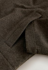 Hnedá vlnená tkanina s jemnou textúrou a elegantným dizajnom. Má plochú kapsu s čistým povrchom, čo zdôrazňuje minimalistický štýl.