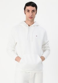Sweat-shirt à capuche blanche avec une poche avant, fabriqué en coton doux. Présente un petit logo sur la poitrine et des poignets côtelés.