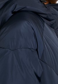 Navyblauwe pufferjas van dichtbij; beschikt over een gladde, glanzende stof, geborduurd patroon, ingetogen capuchon en verstelbare trekkoorddetail.