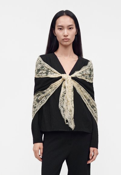 Femme aux longs cheveux noirs porte un top noir à col en V et un pantalon, accessoirisés d’un châle en dentelle écru noué en grand nœud à l'avant.