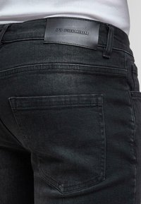 Lähedalt vaadet mustade denimite tagsipoolest, kus on nähtav tagatasku ja naha brändi silt, millel on tekst "2Y PREMIUM."