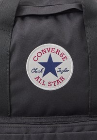 Borsa in tessuto nero con una patch circolare bianca con una stella blu, testo rosso con la scritta "CONVERSE ALL STAR" e "Chuck Taylor". Superficie testurizzata.