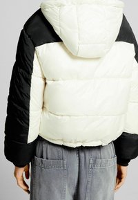 Podpalana kurtka z czarno-kremowym wzorem, z kapturem, pikowaną fakturą i elastycznymi mankietami. Noszona na szarych spodniach z kieszeniami.