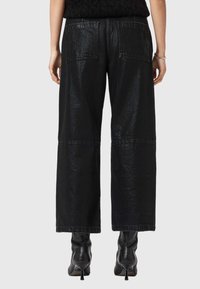 Pantaloni in denim neri a gamba larga con una finitura lucida, dotati di due tasche posteriori e un dettaglio cucito vicino all'orlo. Indossati con stivali neri con tacco.