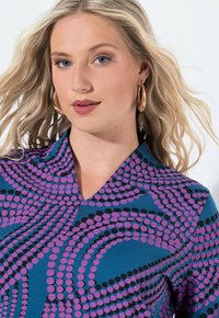 Teal V-Ausschnittoberteil mit einem auffälligen Muster aus lila und schwarzen Punkten, kombiniert mit fließenden Wirbeln. Glatte Textur, taillierte Passform und kurze Ärmel.