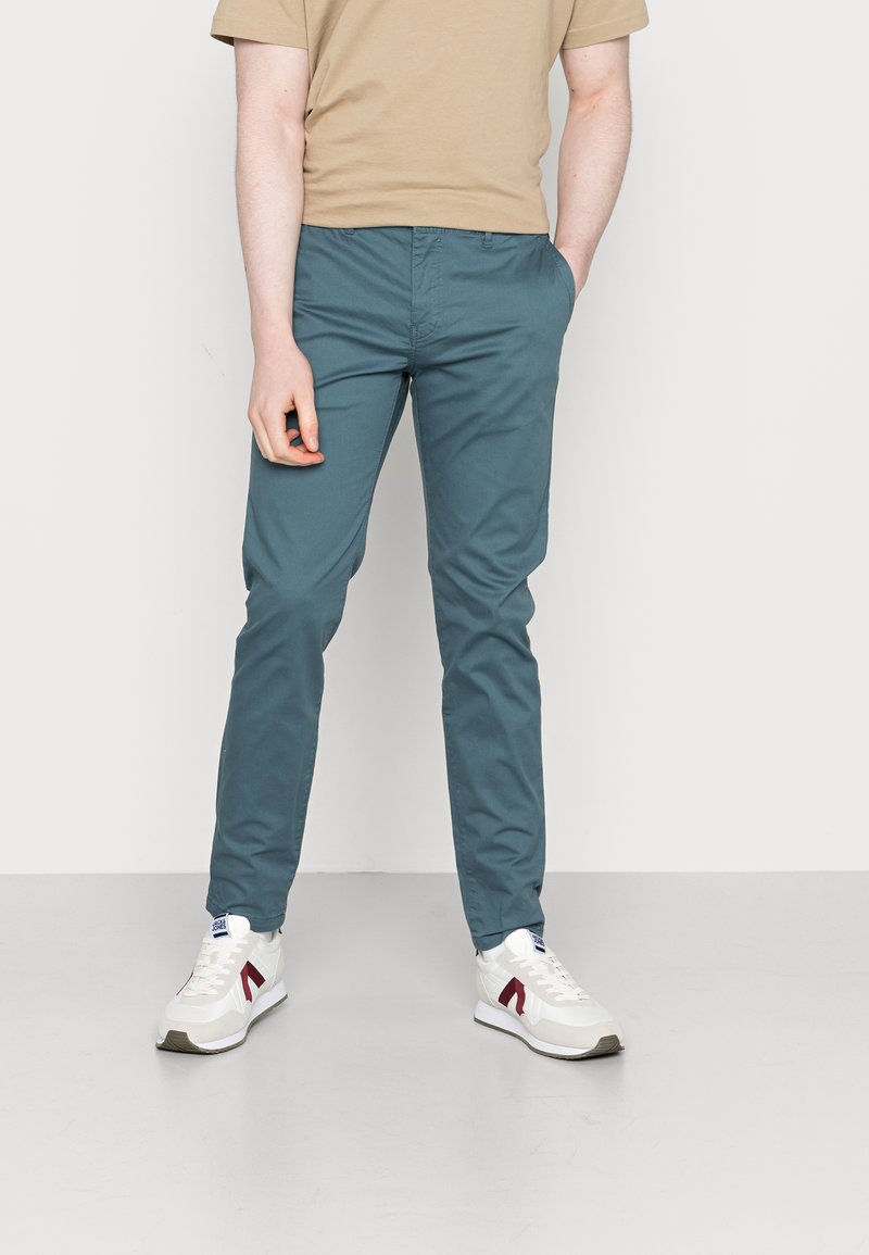 Chino teal ajusté, avec des coutures piquées, des poches latérales et arrière, associé à des baskets blanches et un t-shirt beige.