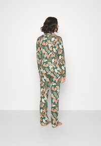 Grünes Pyjama-Set mit Blattmustern in Orange und Creme, lässig geschnittenes Oberteil und Hose mit weichem Material. Barfußmodell von hinten gezeigt.