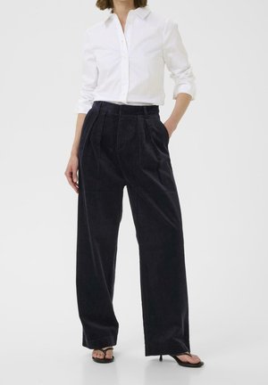 Vrouw draagt een wit overhemd met knopen, ingestopt in een zwarte, wijde, hooggetailleerde broek van ribfluweel, gecombineerd met zwarte sandalen met hak.