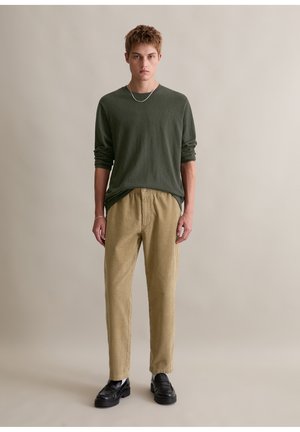 Olijfgroene longsleeve shirt gecombineerd met beige corduroy broek. Model draagt zwarte instappers. Eenvoudig, ontspannen ontwerp.