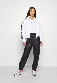 Veste bomber blanche avec des rayures latérales noires, associée à un pantalon en simili cuir noir et des baskets colorées. Le mannequin se tient contre un fond uni.