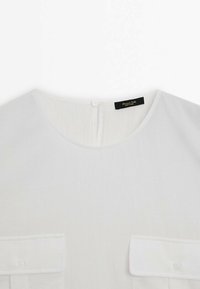 Blouse blanche à col rond, fermeture par boutons au dos, et deux poches avant à rabat avec boutons.