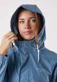 Blå vattentät jacka med huva, silverfärgade detaljer, dragsko och en logotyp på bröstet. Slät, glänsande yta.