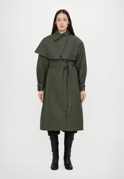 Trench coat verde oliva con colletto, vita cintata e sovrapposizione a mantella. Maniche lunghe con polsini; abbinato a stivali neri fino al ginocchio.
