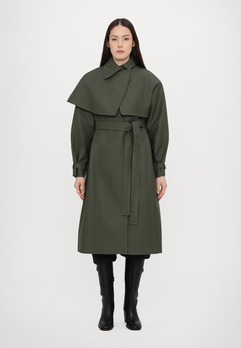 Ελαιόχρωμο παλτό με γιακά, ζώνη στη μέση και κάπα. Μακριά μανίκια με μανσέτες; συνδυασμένο με μαύρες μπότες μέχρι το γόνατο.