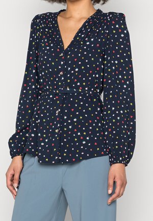 Kvinde iført en navyblå bluse med små flerfarvede blomsterprint, knapper foran, lange puffede ærmer og lyseblå bukser.