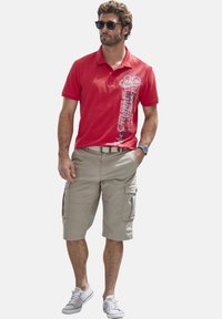 Rotes Poloshirt mit grafischem Druck, kombiniert mit beigen Cargo-Shorts und weißen Turnschuhen. Modell trägt Sonnenbrille und eine Uhr.