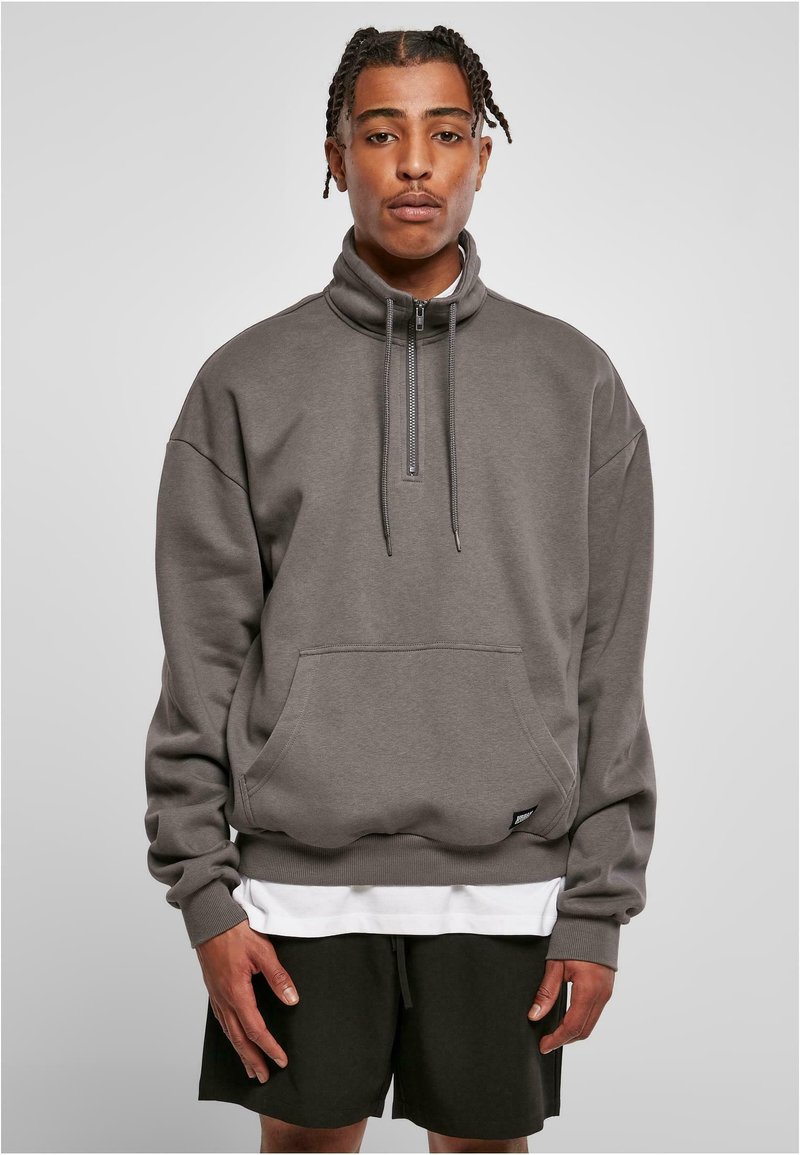 Grå hettegenser med glidelås, høy krage, oversized passform, frontlomme og ribbestrikkede mansjetter. Kombinert med mørke shorts.
