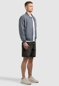 Homme debout portant une veste bomber bleu clair, un t-shirt blanc, un short noir et des baskets crème devant un fond gris uni.