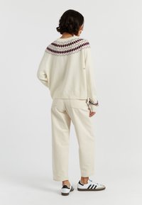 Maglione di maglia crema con un motivo geometrico maroon e grigio sulla spalla, abbinato a pantaloni beige chiaro e sneakers bianco e nere.