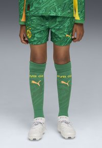 Equipo de fútbol verde que incluye una camiseta con un diseño, pantalones cortos con un logo, y medias altas a juego con detalles amarillos. Botas blancas completan el conjunto.