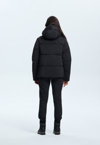 Schwarze Steppjacke mit Kapuze, ausgestattet mit einem gesteppten Design und dreieckigen Nähten. Getragen mit schwarzen Hosen und Stiefeln.