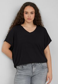 Zwarte V-hals t-shirt met korte mouwen, relaxte pasvorm en zachte textuur; gecombineerd met lichtgrijze jeans. Achtergrond is neutraal grijs.