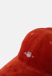 Ruskean värinen korduroykenttä, jossa on brodeerattu logo, jonka ylle on ommeltu viivoja kilpimuotoisen designin ylle. Pinnassa on selvästi erottuva, uritettu rakenne.