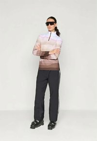 Naine, kes kannab suuri musti prille, gradient-pruuni ja valgega zip-up pusa, musti pükse külgtriipudega ja musti kingi, seismas käed ristus.