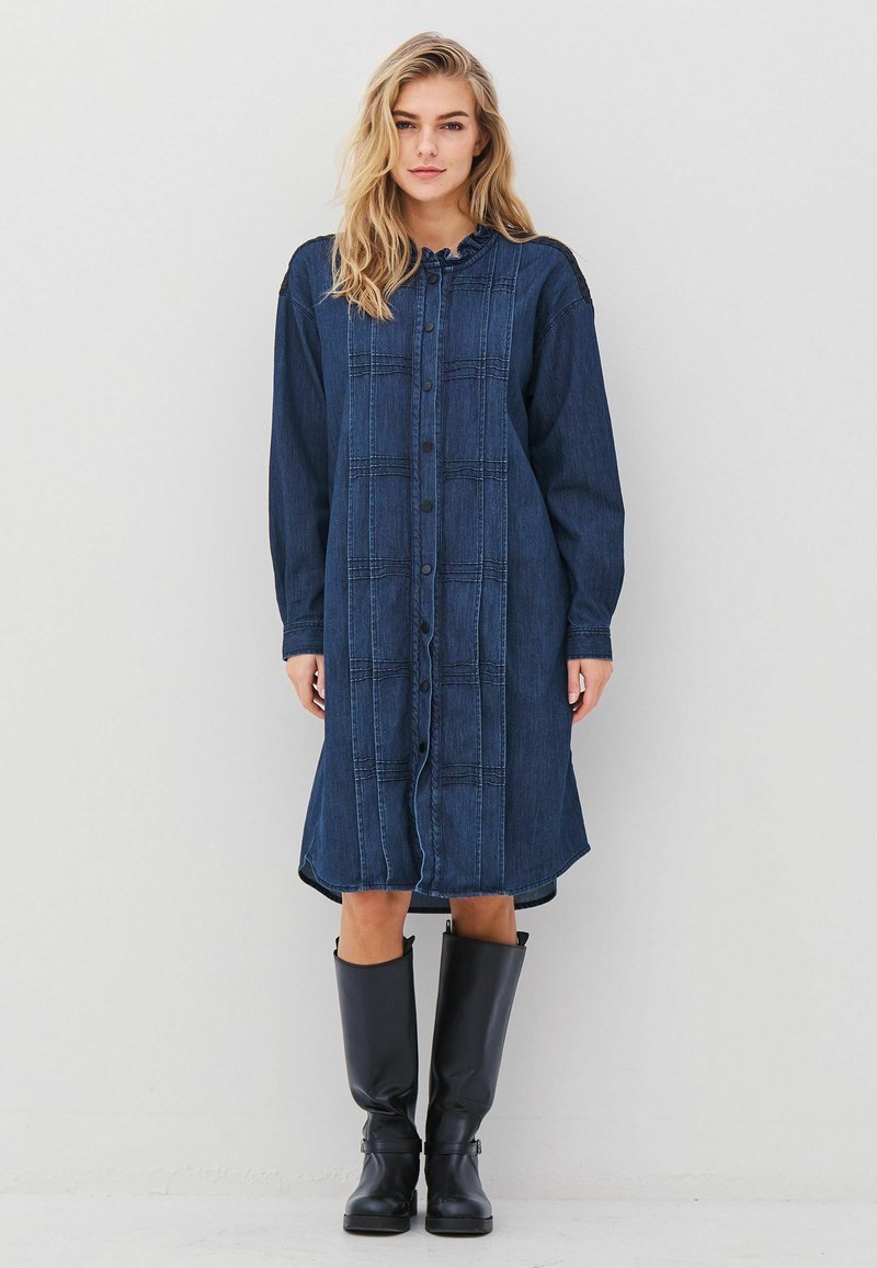 Traper haljina s opuštenim krojem, tamnoplave boje, zatvaranje na dugmiće sprijeda, volanasti izrez i duge rukave, uparena s čizmama do koljena u crnoj boji.