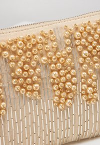 Kopertówka z beżową tkaninową bazą, ozdobiona złotawymi koralikami i wydłużonymi srebrnymi akcentami, tworzącymi teksturowany, wzorzysty design.