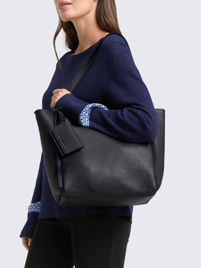 Ženska, ki nosi mornarsko modra vzorčasto jopico in črne hlače ter drži črno usnjeno torbo z ročajem za ramo in pripeto torbico.