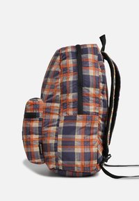 Sac à dos à carreaux multicolores avec des tons orange, violet et crème ; il comprend une poche latérale zippée et des bretelles noires rembourrées.