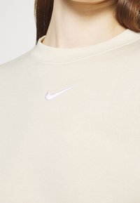 Primo piano di una maglietta Nike beige indossata da una persona, con il logo Nike swoosh bianco ricamato sul petto superiore.