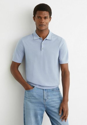 Mand står op ad en hvid væg iført en lyseblå kortærmet polo og blå jeans, med den ene hånd i lommen.