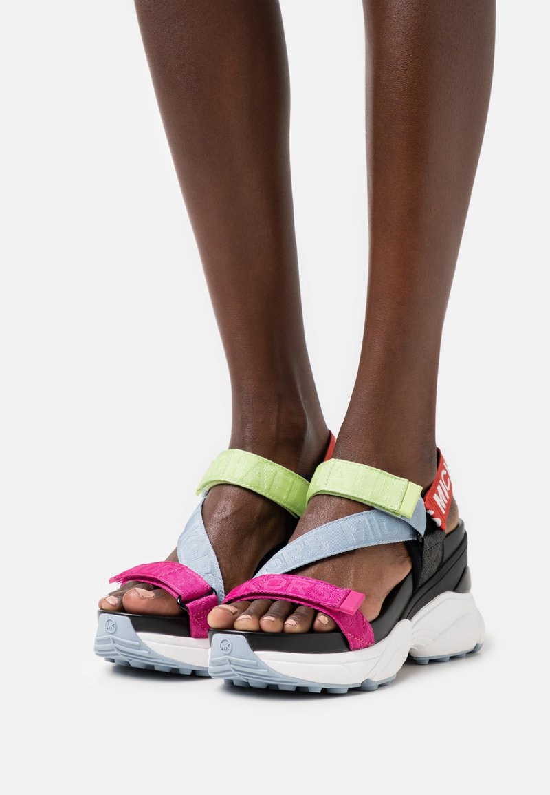 Pieds portant des sandales compensées colorées avec des brides roses, vertes et bleues et des semelles épaisses blanches et grises sur fond blanc.