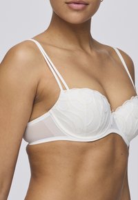 Vitt underwire-bh med broderade vågmönster, tunna justerbara axelband och en genomskinlig sidopanel. Mjuk tyg med en slät yta.