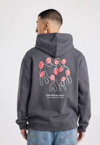 Grijze hoodie met capuchon en trekkoord. Heeft een kleurrijke afbeelding van abstracte figuren op de achterkant met de tekst "SAMEN BOUWEN WE."