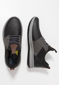 Zapatillas negras y grises con una parte superior lisa, detalles perforados, cordones grises y una suela de goma gruesa. Los acentos incluyen una etiqueta de cuero marrón.
