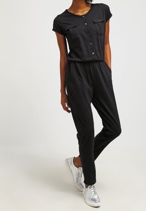 Personne portant une combinaison noire à manches courtes boutonnée avec des poches, associée à des baskets argentées métalliques, debout avec une jambe croisée.