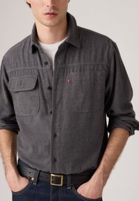 Grijze button-up shirt van textiel, met twee borstzakken en een klein rood logo, gedragen met een zwarte riem en donkere jeans.